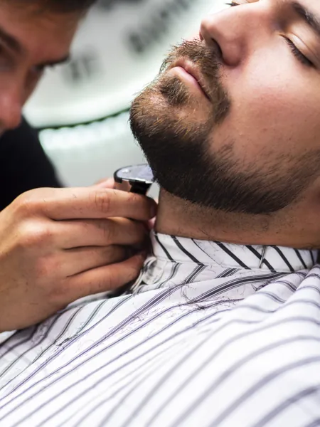 Professional gents salon barber performing beard grooming and shaving service for a male client in a clean, comfortable salon setting.